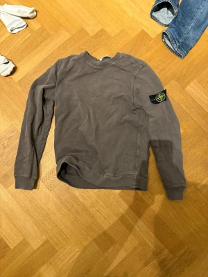 Grå sweatshirt från Stone Island - Säljer en grå långärmad sweatshirt från Stone Island med den klassiska logotypen på vänster ärm. Tröjan har rund hals och ribbade muddar vid ärmslut och nederkant. Tillverkad i mjuk bomull som är skön att ha på sig. Perfekt för dig som gillar streetwear och snygga detaljer.