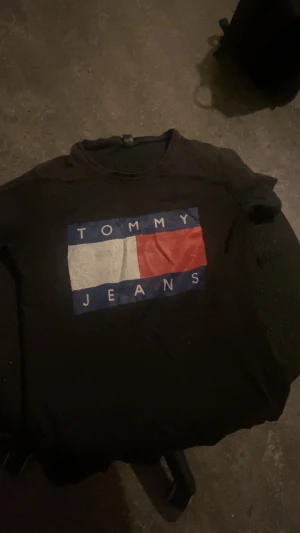 Svart Tommy Jeans t-shirt med tryck size 34 - Svart t-shirt från Tommy Jeans med stort tryck på bröstet i vitt, rött och blått. Klassisk rund hals och korta ärmar. Snygg logga och färgblock ger en cool streetkänsla. Perfekt för dig som gillar ikoniska märken och enkel stil.