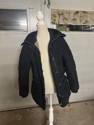 Svart dunjacka från Parajumpers - Svart dunjacka från Parajumpers med huva och broderad patch på ärmen. Jackan har långa ärmar, dragkedja framtill och flera fickor med snygga detaljer. Perfekt för kalla dagar och har en schysst, lite längre passform. Köpt för några år sedan man har ej använt den men än 2 vintrar, lite slitage på armarna men inget som syns. Mycket bra skick !!