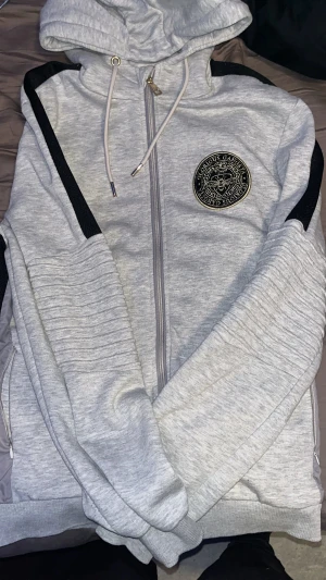 Grå hoodie från Glorious Gangsta - Snygg grå hoodie från Glorious Gangsta med svarta detaljer längs axlar och ärmar. Tröjan har huva med snörning, dragkedja framtill och ett guldigt emblem på bröstet. Mjuk insida i fleece och ribbade muddar för extra komfort.