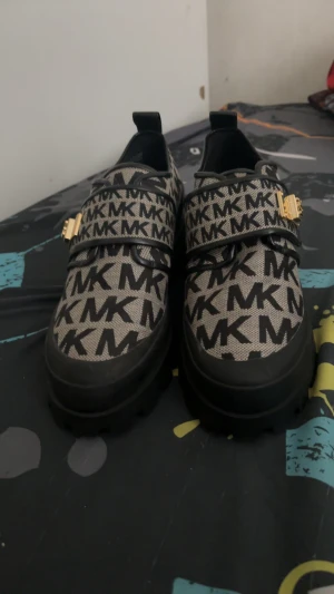 Chunky MK loafers med guldspänne - Säljer ett par chunky loafers från Michael Kors med svart och beige MK-mönster över hela skon. De har bred platt sula, rund tå och ett snyggt guldspänne på ovansidan. Materialet är en mix av textil och skinn, vilket ger en cool kontrast. Perfekt för dig som gillar statement skor.