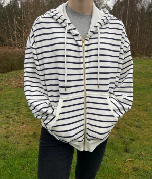 Vit och marinblå randig hoodie - Säljer en oversized hoodie med marinblå ränder på vit botten. Tröjan har huva med snörning och guldfärgade detaljer på snöret, dragkedja framtill och stora fickor. Perfekt för chill dagar och har en riktigt skön passform. Sparsamt använd, så fint skick.