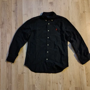 Mörkblå skjorta från Ralph Lauren S - Mörkblå långärmad skjorta från Ralph Lauren med klassisk krage och knäppning framtill. Skjortan har det ikoniska röda broderade Polo-loggan på bröstet och är tillverkad i mjuk bomull. Perfekt för dig som gillar stilrena och tidlösa plagg. Nypris: va 1500