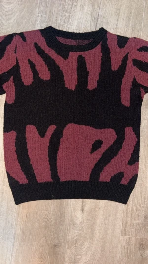 Vinröd och svart stickad tröja - Säljer en cool stickad tröja med vinrött och svart abstrakt mönster. Tröjan har rund hals och långärmade ärmar med ribbade muddar. Perfekt för dig som gillar att sticka ut med grafiska detaljer och mjukt material.