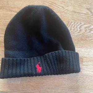 Svart stickad mössa från Polo Ralph Lauren - Svart stickad mössa från Polo Ralph Lauren med klassisk röd broderad logga framtill. Mössan har ribbad kant och är tillverkad i mjukt material som håller dig varm under kyliga dagar. Perfekt accessoar för en clean och stilren look.