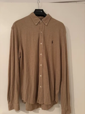 Beige skjorta från Ralph Lauren  - Klassisk beige skjorta från Ralph Lauren i storlek L. Tillverkad i lätt bomullsmaterial med button-down krage, långa ärmar och vita knappar framtill. Diskret broderad logga på bröstet. 