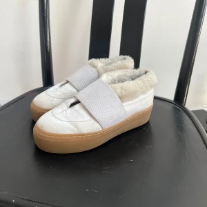 Vita sneakers med fuskpäls från Rodebjer - Unika vita sneakers från Rodebjer med bred elastisk rem över foten och mjuk beige fuskpäls på insidan. Snygg platt gummisula i brunt och rund tå. Perfekt för dig som vill ha något annorlunda och mysigt på fötterna.