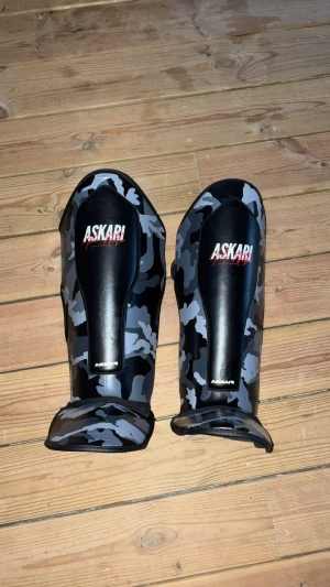 Askari kamouflage benskydd - Säljer ett par svarta och grå kamouflage benskydd från Askari. De har en cool camo-design och är tillverkade i syntetmaterial med vaddering för extra skydd. Bra för sparring, skriv ifall du har frågor eller för mer bilder.