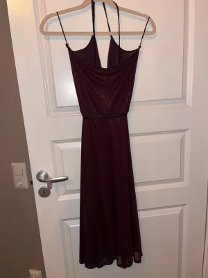 Vinröd glittrig långklänning med knyt - Vinröd långklänning med glittrigt tyg och smala axelband. Klänningen har omlott-drapering framtill, flätade detaljer vid axelband och midja samt ett knytbälte med pärlor. Perfekt för dig som vill sticka ut med en elegant och trendig look.