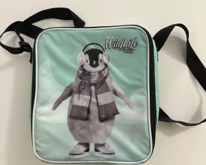 WildLife axelväska med pingvinmotiv - Cool axelväska med mintgrön framsida och ett pingvinmotiv med hörlurar och halsduk. Väskan har svart justerbar axelrem och dragkedja runt hela. Insidan är silverfärgad och isolerad, perfekt för att hålla saker kylda. Trycket är unikt och väskan är fyrkantig i formen.