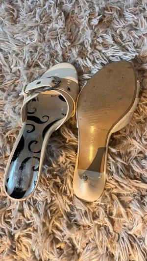 Vita klackade sandaler med spänne - Säljer ett par vita sandaler med hög klack och öppen tå. Skorna har ett brett band över foten med ett snyggt spänne i silver och quiltad detalj. Snyggt mönstrad innersula och stilren design. Perfekta för dig som vill sticka ut lite extra.