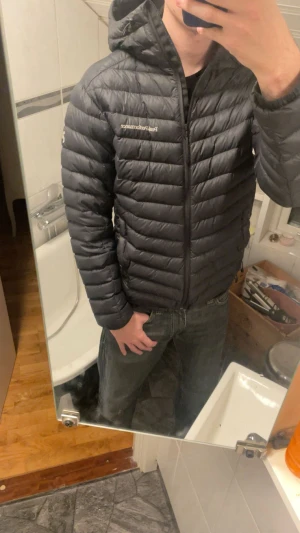 Svart dunjacka Peak Performance S - Svart dunjacka från Peak Performance i storlek S. Jackan är lätt, har huva och dragkedja framtill. Tillverkad i Pertex Quantum-material som ger bra vindskydd och värme. Klassisk pufferstil med horisontella sömmar och diskret logga på bröstet.