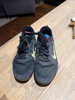 Nike Vapormax Flyknit svarta sneakers - Nike Vapormax Flyknit sneakers i svart med blå detaljer och neongul swoosh. Ovandel i stickad flyknit, svarta snören med rosa tips och blå etikett på plösen. Genomskinlig plattformssula med limegröna inslag. Reflekterande dragflik bak.