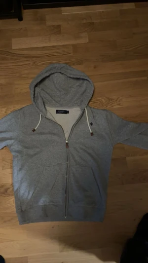 Grå hoodie från Morris med dragkedja - Snygg grå hoodie från Morris med huva och vita snören. Tröjan har dragkedja framtill, två fickor och en liten broderad logga på bröstet. Tillverkad i mjuk bomullsmix som är skön att ha på sig. Perfekt för chill eller när du vill vara casual.