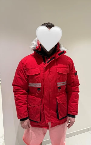 Röd parkas First Slope Expedition - Röd parkas från First Slope Expedition med reflexdetaljer, flera stora fickor och huva med fuskpäls. Jackan har svart innerfoder med stort tryck, justerbar midja och dragkedja framtill. Perfekt för kalla vinterdagar och riktigt snygg streetstil.