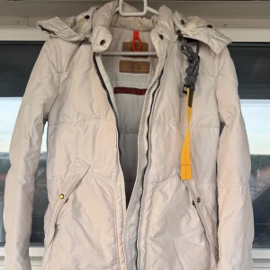 Vit dunjacka från Parajumpers - Säljer en vit dunjacka från Parajumpers med huva och dragkedja. Jackan har långa ärmar, två snedställda fickor med knappar och en gul detalj. 