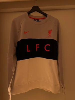 Nike Liverpool LFC sweatshirt grå/svart/vit - Snygg sweatshirt från Nike med Liverpool FC-tryck i rött på bröstet. Tröjan har en bred svart panel över bröstet med LFC-text, grå ovandel och vit nederdel. Rund hals och långa ärmar, perfekt för dig som gillar fotboll och streetwear.