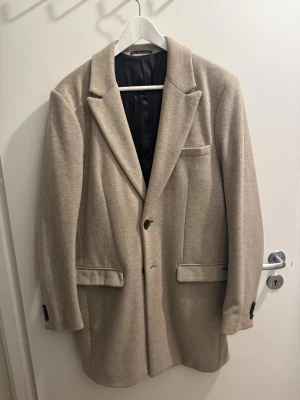 Beige/grå kappa från Scotch & Soda - Stilren beige/grå kappa från Scotch & Soda med klassisk passform, två svarta knappar framtill och fyra dekorativa knappar vid ärmslutet. Kappa har tre fickor,2 med lock och är fodrad med marinblått tyg. Perfekt för dig som gillar en clean och tidlös look.