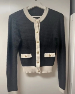 Svart och vit kofta med guldknappar - Supersnygg svart kofta med vit kontrastkant och guldiga knappar framtill. Koftan har ribbad struktur, långa ärmar och två dekorativa fickor med vita lock och guldknapp. Croppad passform som ger en trendig look, perfekt att styla med jeans.