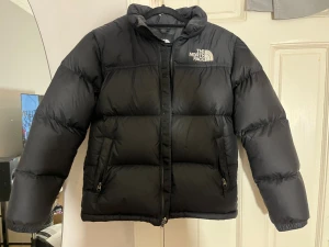 Svart pufferjacka från The North Face - Säljer en svart pufferjacka från The North Face med broderad logga på bröstet och ryggen. Jackan har hög krage, dragkedja framtill och två fickor med dragkedja. Perfekt för kalla dagar och riktigt snygg streetstil.