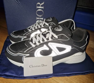 Svarta sneakers från Christian Dior - Säljer ett par svarta sneakers från Christian Dior med vit kontrastdetalj och stora CD-loggor på sidorna. Skorna har snörning, rund tå och en platt sula. Materialet är en mix av syntet och mesh för en sportig vibe. Perfekta för dig som gillar exklusiva märken och streetstyle.