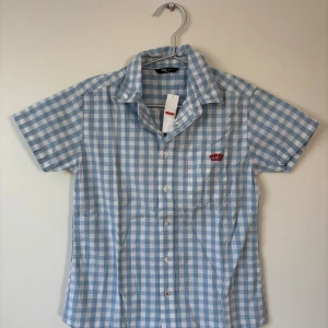 Kortärmad skjorta - 134/140 - Kortärmad skjorta från  i ljusblått och vitt rutigt mönster. Klassisk krage, knäppning framtill och liten bröstficka med röd brodyr. Skjortan är i tunn bomull, 