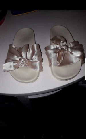 Beiga sandaler med satinrosett - Snygga beiga sandaler med bred rem i glansigt satinmaterial och stora rosetter framtill. Platt sula och öppen tå gör dem perfekta för varma dagar. Superfina att matcha med somriga outfits för en söt och trendig look.