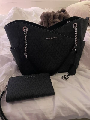 Svart Michael Kors axelväska + plånbok - Snygg svart axelväska från Michael Kors med silverfärgade kedjedetaljer och MK-logga. Väskan har ett diskret mönster med MK-bokstäver och flera fack. Tillhörande plånbok i samma stil och material ingår. Perfekt för dig som gillar stilrena accessoarer.