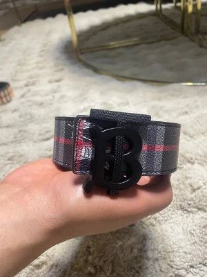 Rutig Burberry bälte med TB-spänne - Snyggt bälte från Burberry med klassiskt rutigt mönster i svart, grått och rött. Bältet har ett stort svart TB-spänne och är tillverkat i skinn. Perfekt accessoar för att lyfta din outfit med en lyxig touch.