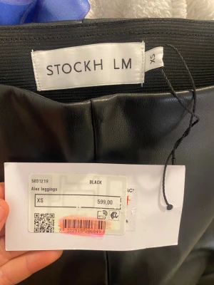Svarta leggings i skinnimitation XS - Snygga svarta leggings från Stockholm LM i skinnimitation. Modellen heter Alex och har en hög midja med elastisk linning. Byxorna är tighta och har en stilren look som passar till många olika outfits. Perfekt för dig som gillar trendiga och bekväma byxor. Original pris 600 kr. ALDRIG andvända