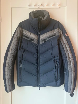 Blå Moncler Grenoble dunjacka - Snygg Moncler Grenoble dunjacka i mörkblått och grått med coola detaljer. Jackan har hög krage, dragkedjor framtill och på bröstet, samt Moncler-logga på ärmen. Insidan är fodrad i silver och har smart ficka för glasögon och putsduk. Perfekt för kalla dagar.