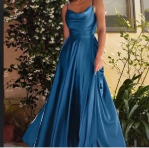 Blå balklänning i satin med slits - Säljer en elegant blå långklänning i glansigt satinmaterial. Klänningen har smala axelband, öppen rygg och en snygg slits på sidan. Den har en mjukt draperad urringning och knytband vid axlarna för extra detalj. Perfekt för dig som vill sticka ut.