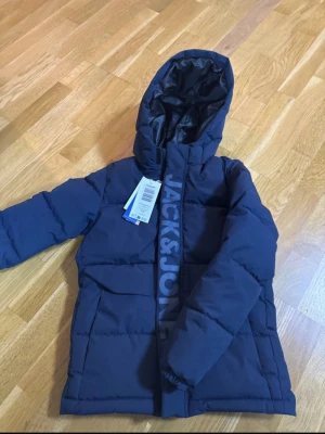 Mörkblå pufferjacka från Jack & Jones - Snygg mörkblå pufferjacka från Jack & Jones med huva och stora fickor framtill. Jackan har tydlig logga längs dragkedjan och är vadderad för extra värme. Perfekt för kalla dagar och har en modern, enkel look. Helt ny med prislapp. 