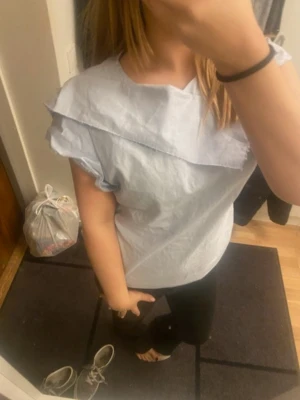 Ljusblå randig blus med volangdetalj - Säljer en ljusblå/vit randig blus med korta ärmar och en snygg volangdetalj över axlarna. Blusen har rund halsringning och dragkedja i ryggen. Perfekt för dig som gillar en stilren men ändå unik look.