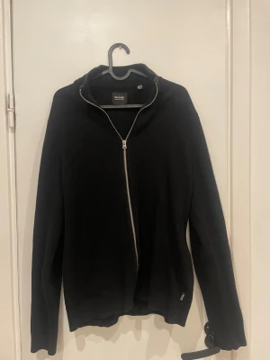 Svart stickad kofta från Only & Sons - Svart stickad kofta från Only & Sons med hel dragkedja framtill och ribbad struktur. Tröjan har långärmad design och en stilren, enkel look som passar perfekt till jeans eller chinos. Snygg ståkrage och diskret logga vid nederkanten.