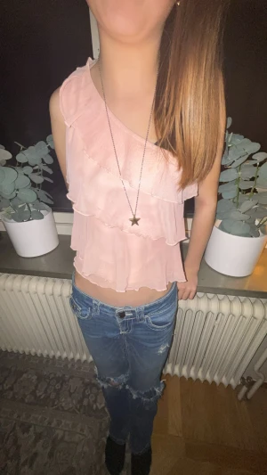 Ljusrosa volang topp oneshoulder - Säljer en ljusrosa oneshoulder topp med flera lager volanger och omlott-design. Toppen är croppad och har en luftig, lätt känsla som passar perfekt till jeans. Materialet är tunt och ger ett snyggt fall över kroppen. Perfekt för dig som gillar romantiska detaljer och unik stil.