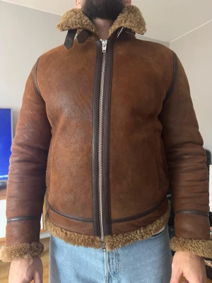 Brun lamm pälsfodrad skinnjacka med dragkedja - Säljer en brun skinnjacka med pälsfoder och bred krage. Jackan har detaljer i mörkare brunt läder, synliga sömmar och grov dragkedja framtill. Pälsen syns vid krage, ärmslut och nederkant. Perfekt för kalla dagar och ger en riktigt snygg vintage vibe.