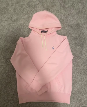 Polo Ralph Lauren hoodie - Snygg ljusrosa hoodie från Polo Ralph Lauren med klassisk huva och snörning. Tröjan har en broderad blå logga på bröstet och en stor ficka framtill. Mjuk bomullskvalitet och normal passform, perfekt för lugna dagar. Den finns en liten vit fläck där bak men som inte syns ordentligt verkligheten. Kvitto finns såklart 