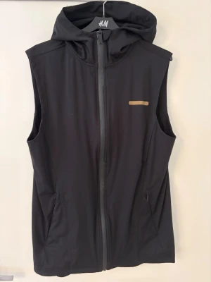 Svart träningsväst med huva från A–Z - Sportig svart väst från A–Z i storlek M, perfekt för träning. Västen har huva, hel dragkedja framtill och två sidofickor med dragkedja. Snyggt minimalistiskt tryck på bröstet och ryggen. Materialet är syntet och designen är clean och funktionell.