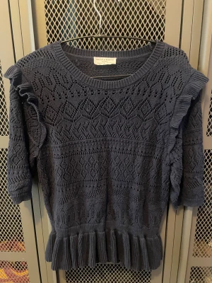 Mörkblå stickad tröja Holly & Whyte - Supersöt mörkblå stickad tröja från Holly & Whyte med snyggt hålmönster och volangdetaljer på axlar och nederkant. Rund halsringning och trekvartsärm ger en chill vibe. Perfekt för dig som gillar unika detaljer och vill sticka ut lite extra.