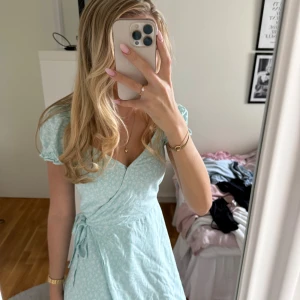 Ljusblå omlottklänning från Hollister - Ljusblå omlottklänning från Hollister med små vita blommor, kort ärm och knyt i midjan. V-ringning och lätt volang vid ärmslut. Har gula fläckar på insidan av ärmarna men det syns inte utåt då det är på insidan av tyget.