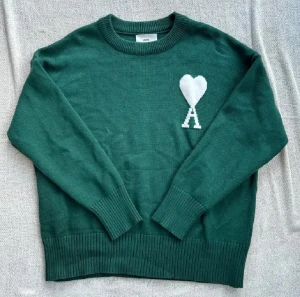 Grön stickad tröja från Ami - Snygg mörkgrön stickad tröja från Ami med vit hjärt- och A-logga på bröstet. Tröjan har rund hals, ribbade muddar och är tillverkad i mjuk ull. Perfekt för dig som gillar stilrena detaljer och vill ha något som sticker ut lite extra.