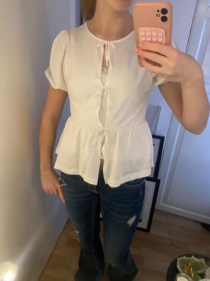 Vit blus med rosetter och puffärm - Supersöt vit blus med korta puffärmar och tre knyt framtill. Blusen har en luftig och flowy passform med volang nedtill. Materialet känns lätt och skönt mot huden. Använt en gång.