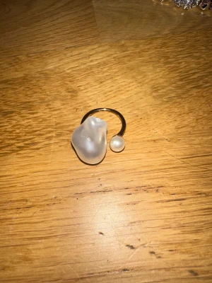 Guldig ring med oregelbunden pärldetalj - Unik guldig ring med en stor, oregelbunden pärlliknande detalj och en mindre pärla. Ringen har en öppen design och cool, modern vibe. Perfekt för dig som gillar statement-smycken och vill sticka ut med något annorlunda.