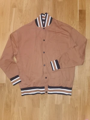 NY Brun bomberjacka från ASOS DESIGN, storlek   - Snygg brun bomberjacka från ASOS DESIGN med svarta, vita och bruna ränder på krage, ärmslut och midja. Jackan har knappar framtill och två fickor. Tillverkad i mjuk bomullsmix och har en klassisk, sportig look. Jackan är oanvänd, alltså Ny då storleken vart för liten. Storlek S