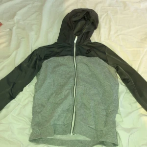 Grå och svart Nike vindjacka med huva - Nike vindjacka i grått och svart med huva och hel dragkedja framtill. Jackan har långärmade svarta ärmar och grå kropp, samt Nike-logga på bröstet och ryggen. Perfekt för chill eller träning, snygg och sportig look.