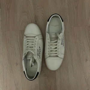 Saint Laurent vita sneakers i skinn - Snygga vita sneakers från Saint Laurent med svart detalj på hälen och handskriven logga på sidan. Skorna har rund tå, platt sula och klassisk snörning. Tillverkade i skinn för en lyxig känsla och stilren look.