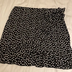 Svart blommig omlottkjol med volang - Supersöt svart kort omlottkjol med vita blommor och volangkant. Kjolen knyts i sidan och har en flowy känsla, perfekt för en trendig look. Lätt och luftig, passar perfekt till sneakers eller sandaler.