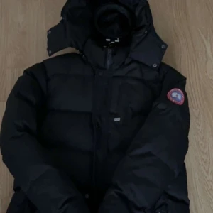 Svart dunjacka från Canada Goose - Säljer en svart dunjacka från Canada Goose med huva och klassisk logga på ärmen. Jackan har en puffad design, dragkedja framtill och flera fickor. Perfekt för kalla dagar och har en riktigt schysst streetstil.
