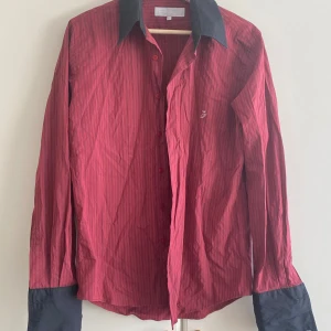 Röd- och svart randig skjorta Poca Loco - Snygg röd skjorta från Poca Loco med svarta tunna ränder, svart krage och svarta muddar. Klassisk passform med knappar framtill och diskret logga på bröstet. Perfekt för dig som vill sticka ut lite extra med kontrastdetaljer.